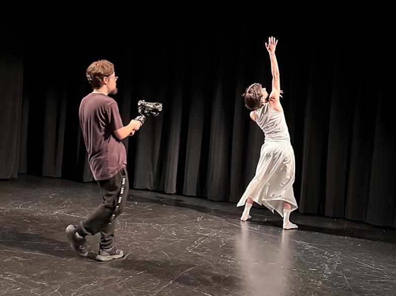 Un jeune filme une performance de danse sur scène : une danseuse vêtue de blanc lève un bras vers le haut, sous un éclairage de théâtre doux, devant un rideau noir.