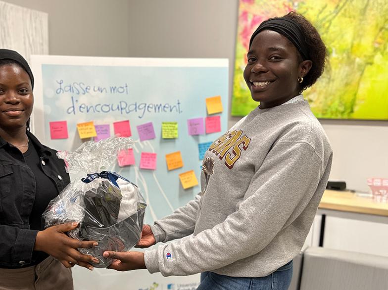 Deux étudiantes souriantes tiennent ensemble un panier-cadeau lors de la Journée du bienêtre à l’USB, devant un tableau rempli de mots d’encouragement colorés.