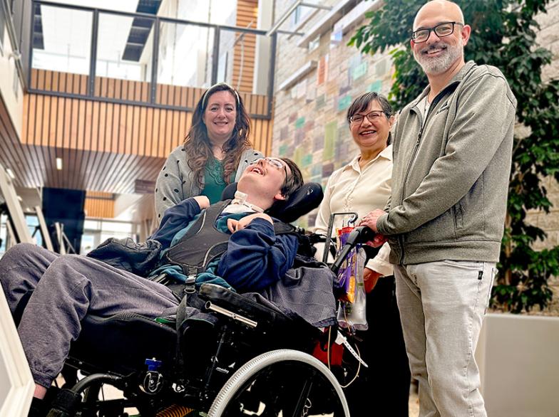 Un jeune homme en fauteuil roulant sourit, entouré de trois adultes dans l’Atrium de l’USB, devant une œuvre exposée; ambiance chaleureuse et inclusive.