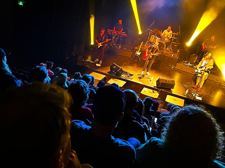 Vue depuis les gradins d’une salle de spectacle où un public attentif regarde le groupe La Chicane performer sous des éclairages jaunes sur scène.