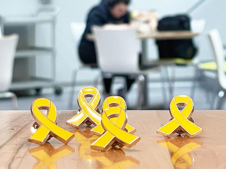 Gros plan sur des épinglettes en forme de ruban jaune posées sur une table, avec des étudiantes et étudiants flous en arrière-plan.