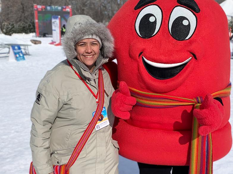 Une femme métisse en manteau d'hiver et un sac au patron d'une ceinture fléchée, accompagnée d'une personne costumée en tuque rouge.