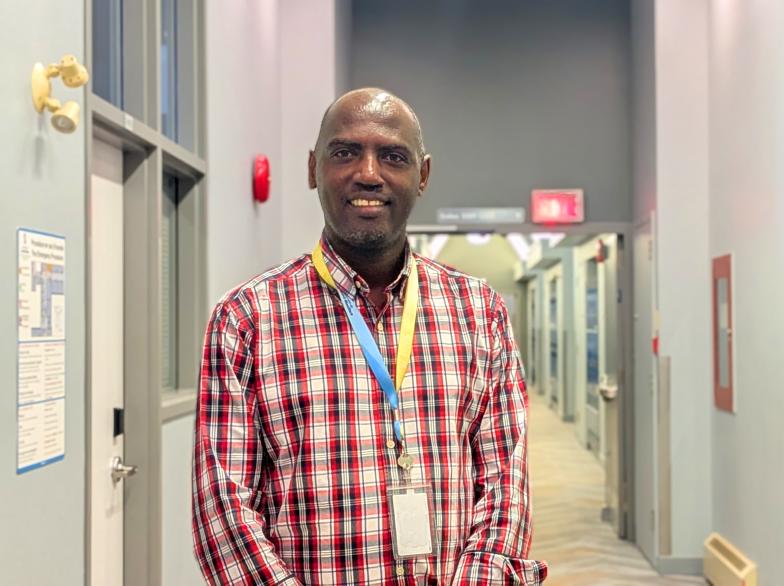 Jean Pacifique Niyongabo, debout dans un couloir de l'université, sourit vers l'objectif de la caméra.
