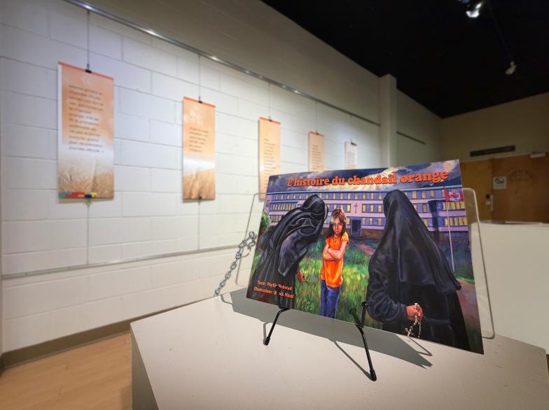 Le livre « L'histoire du chandail orange », placé sur un support au milieu d'une galerie d'art, des panneaux orange fixés au mur en arrière-plan.