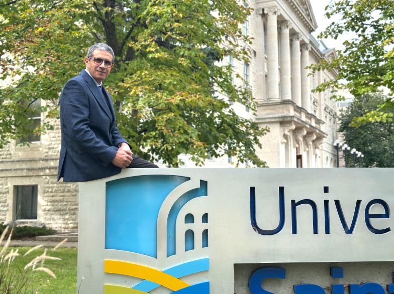 Brahim El Fadil, doyen de la Faculté des arts et Faculté des sciences, assis sur le monument de l'Université de Saint-Boniface, le bâtiment en arrière-fond.