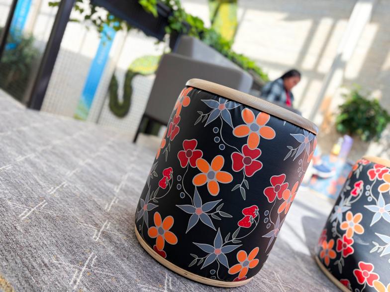 Un siège ressemblant à un tambour peint avec des fleurs, dans une espace éclairé avec verdure, une étudiante floutée en arrière-fond.