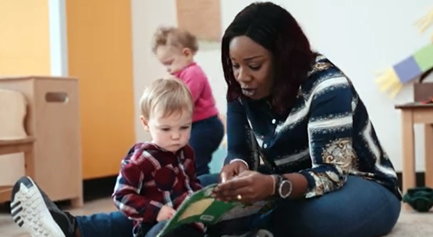 Une femme assise par terre regarde un livre avec un tout-petit à sa droite. Un autre enfant en bas âge est debout derrière eux. 