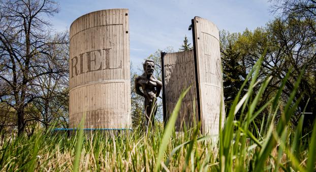 Statue de Louis Riel placée entre deux grandes structures de béton courbées gravées du nom "RIEL", entourée d’arbres et vue à travers l’herbe haute au premier plan, sous un ciel bleu.