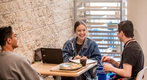 Une étudiante souriante, assise avec un ordinateur portable et un cahier à une table de cafétéria, discute avec un camarade devant un repas.