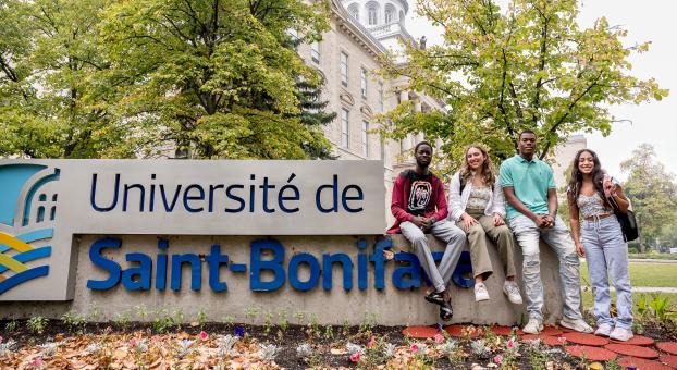 Quatre étudiants assis sur le muret signalétique devant l’USB, des arbres feuillus et la coupole du bâtiment sont visibles en arrière-plan.