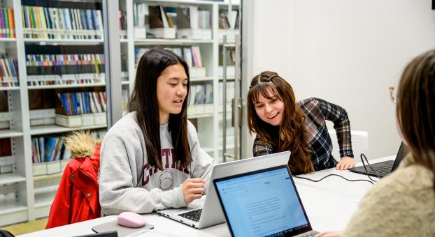 Deux étudiantes souriantes travaillent ensemble sur un ordinateur portable dans une salle de bibliothèque.