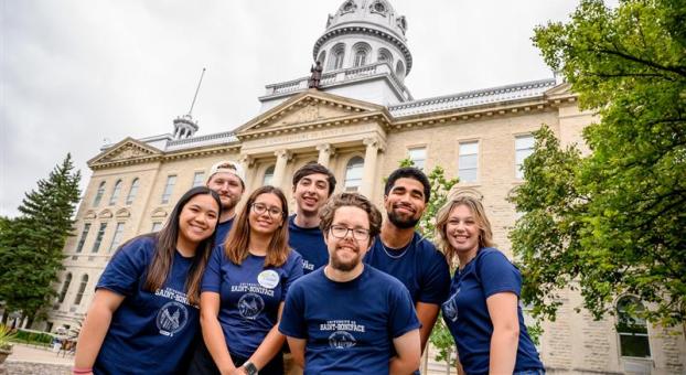 Sept personnes souriantes en t-shirt bleu regroupées devant le bâtiment de l'Université de Saint-Boniface.