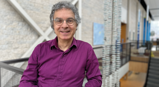 Portrait de David Alper, souriant, portant une chemise mauve, debout dans un espace intérieur lumineux de l’Université de Saint-Boniface.