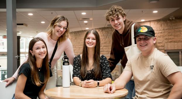  Cinq personnes réunies autour d’une table sourient et regardent l’objectif de la caméra dans un espace intérieur lumineux.