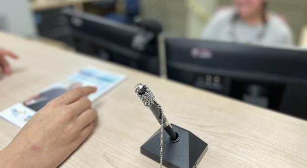 Un stylo à chaîne fixé sur un socle repose sur un comptoir, une main tient un dépliant, et en arrière‑plan flou, une personne à un poste informatique. 