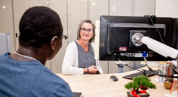 Une dame souriante accueille à son poste de travail un étudiant.