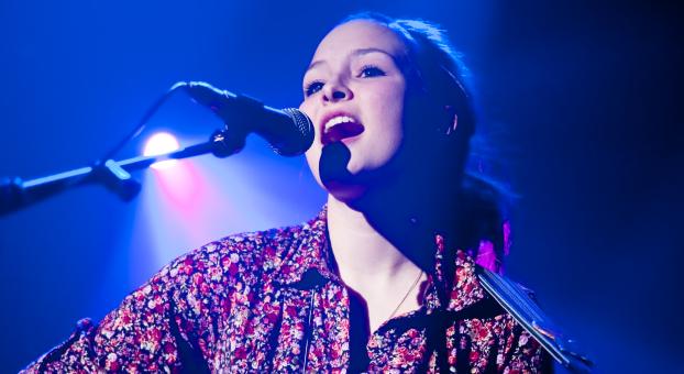 Une jeune guitariste chante sur scène, illuminée d'une lumière bleue.