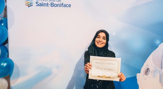 Une étudiante brandit fièrement son certificat devant un écran aux couleurs de l’université. 