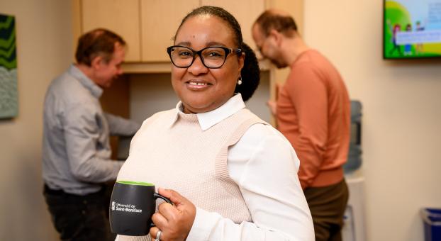 Une femme tenant une tasse de café regarde l'objectif, des collègues derrière elle près d'une imprimante.
