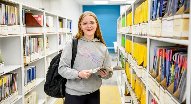 Une jeune femme aux long cheveux roux et vêtue de gris est debout entre deux rayons de bibliothèque.