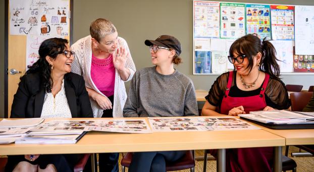 Une enseignante discute avec des personnes apprenantes dans une salle de classe, des cahiers sont étalés sur la table.