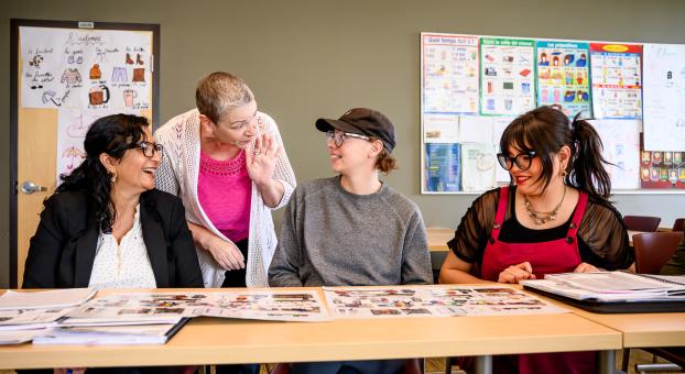 Une enseignante discute avec des personnes apprenantes dans une salle de classe, des cahiers sont étalés sur la table.