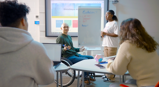 Salle de classe collaborative où un étudiant en fauteuil roulant participe à un travail d'équipe devant un tableau, en présence d'un étudiant et de deux étudiantes.  