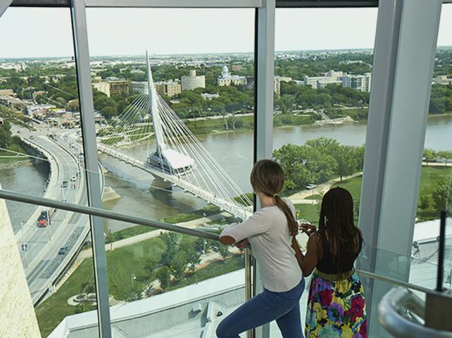 Deux femmes monte l'escalier du Musée canadien pour les droits de la personne, regardant à travers une baie vitrée le pont Esplanade Riel qui enjambe la rivière Rouge à Winnipeg.