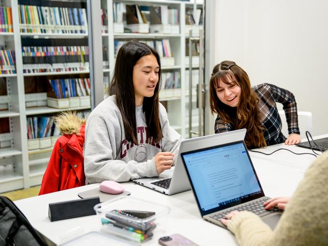 Trois personnes assises à une table consultent des ordinateurs. L’une d’elles est penchée vers sa collègue et semble rire.