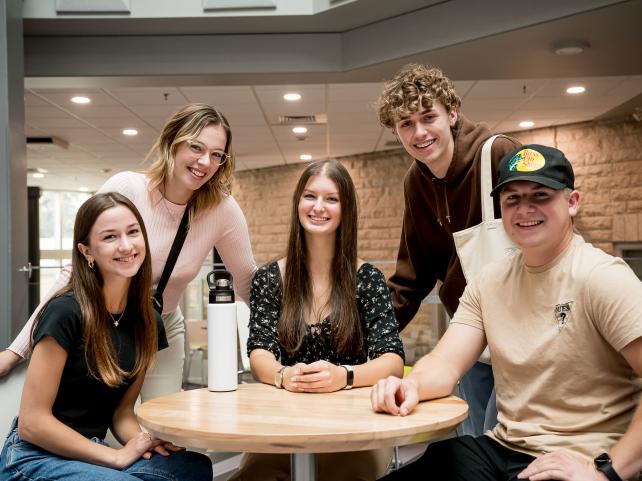  Cinq personnes réunies autour d’une table sourient et regardent l’objectif de la caméra dans un espace intérieur lumineux.