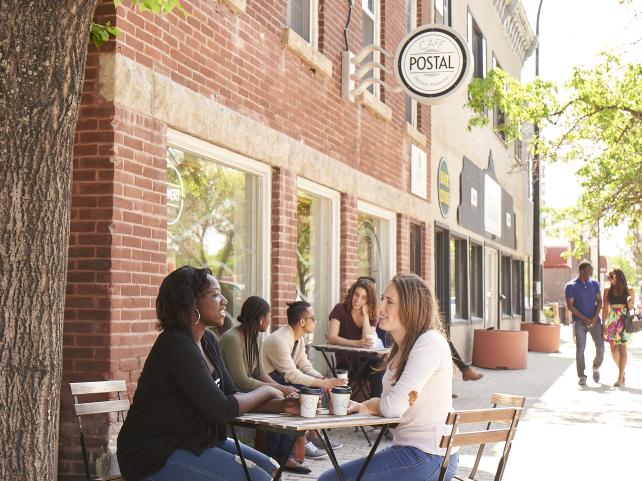 Deux étudiantes souriantes discutent à une table en extérieur devant un café par une journée ensoleillée.