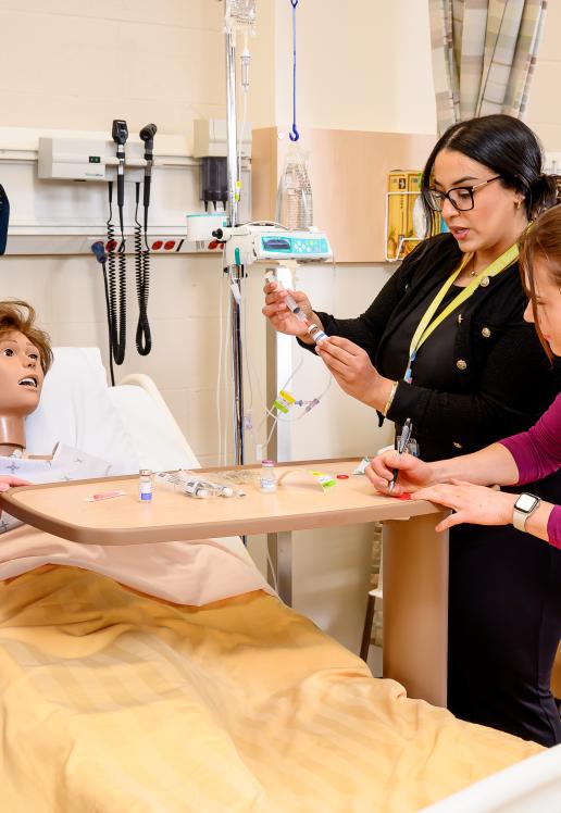 Une professeure démontre une technique de seringue à quelques personnes étudiantes autour d’un lit d’hôpital dans lequel est couché un mannequin.