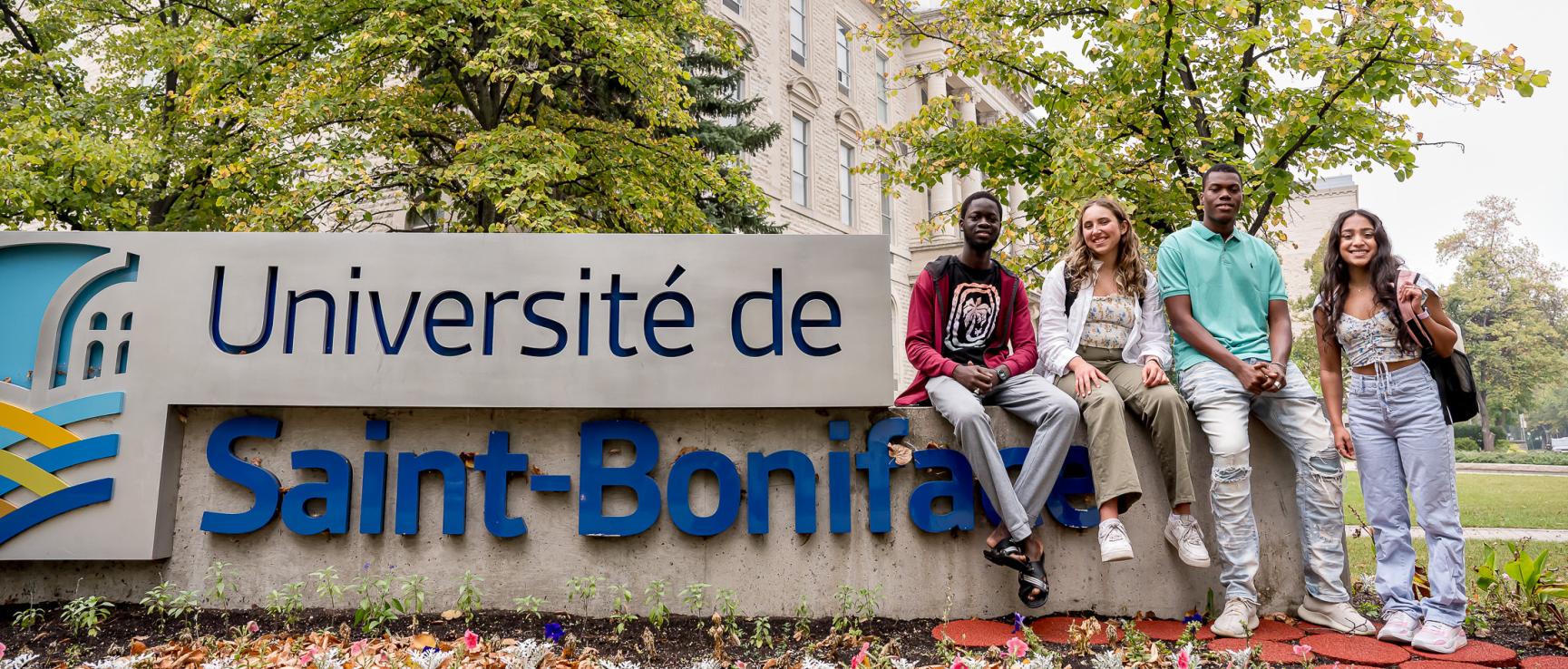 Quatre étudiants assis sur le muret signalétique devant l’USB, des arbres feuillus et la coupole du bâtiment sont visibles en arrière-plan.