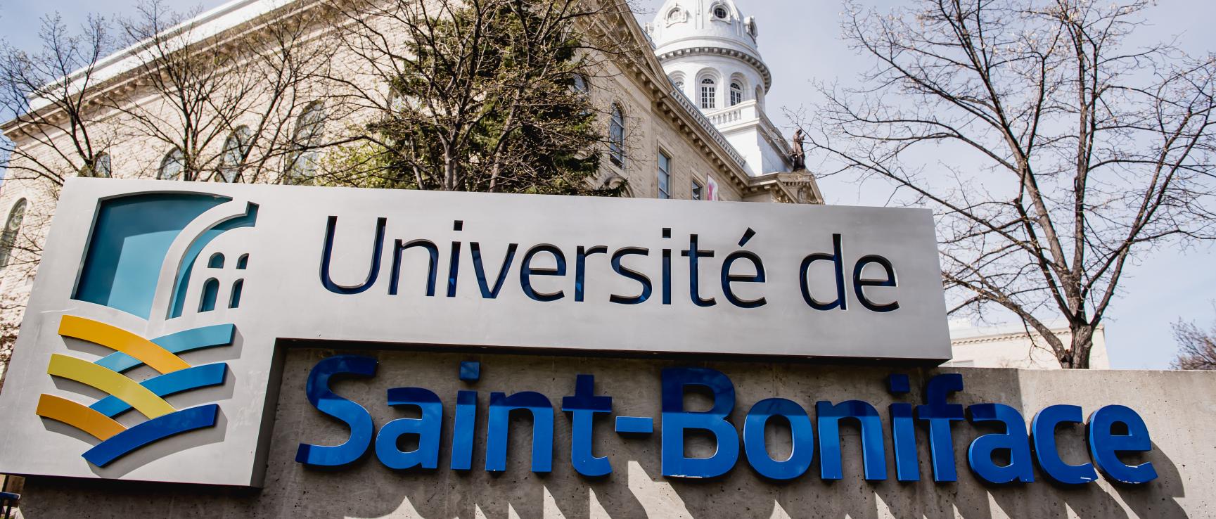 Monument d'entrée de l'Université de Saint-Boniface devant le bâtiment un jour de printemps, des arbres sont dépouillés de leurs feuilles.