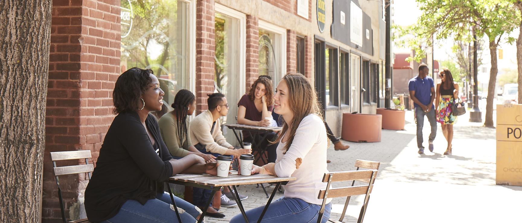 Deux étudiantes souriantes discutent à une table en extérieur devant un café par une journée ensoleillée.