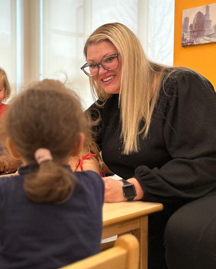 Une femme aux longs cheveux blonds portant des lunettes sourit en se penchant vers une jeune personne assise vue de dos. 