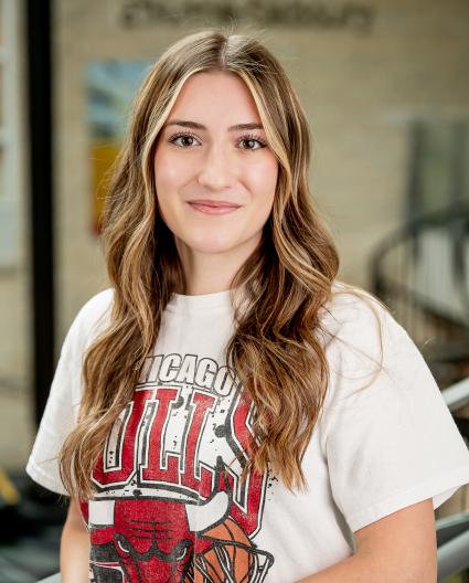 Une jeune femme aux cheveux châtains longs et ondulés sourit à la caméra. Elle porte un t-shirt blanc de l’équipe de basketball des Chicago Bulls.