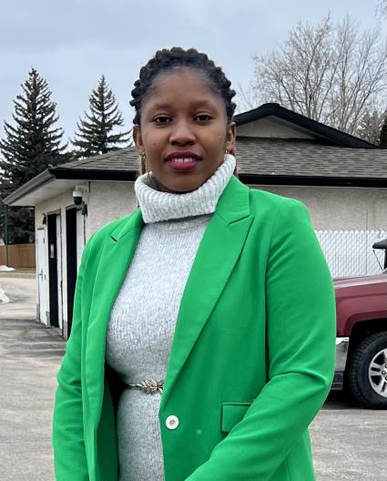 Une femme portant une robe gris pâle et un blazer vert regarde l’objectif. Elle est dehors, et il y a une maison en arrière-plan. 