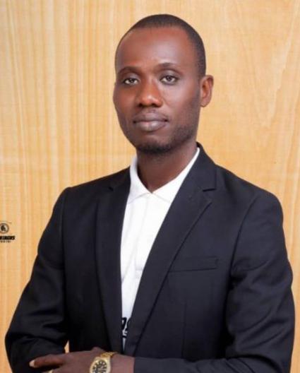 Un homme aux cheveux noirs très courts, portant une chemise blanche et un veston noir, regarde vers l’objectif les bras croisés. 
