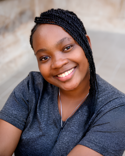 Une femme aux cheveux noirs tressés, vêtue de bleu, penche la tête tout en souriant vers l’objectif