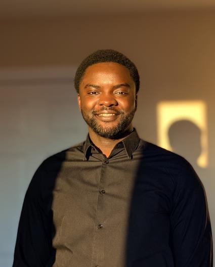 Un homme souriant aux cheveux noirs, vêtu d’une chemise brun foncé, se tient debout dans un rayon de soleil, projetant une ombre derrière lui.