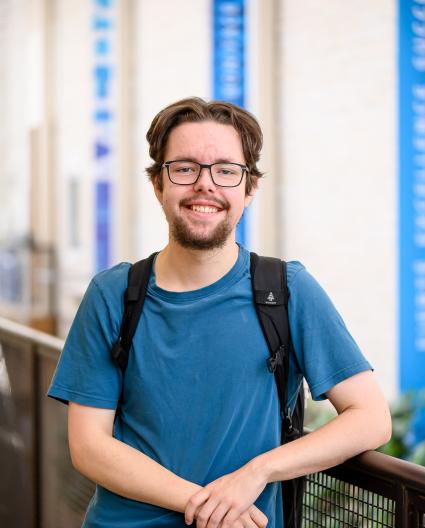 Un jeune homme portant des lunettes, un t-shirt bleu et un sac à dos sourit vers l’objectif. Il s’appuie sur une balustrade.