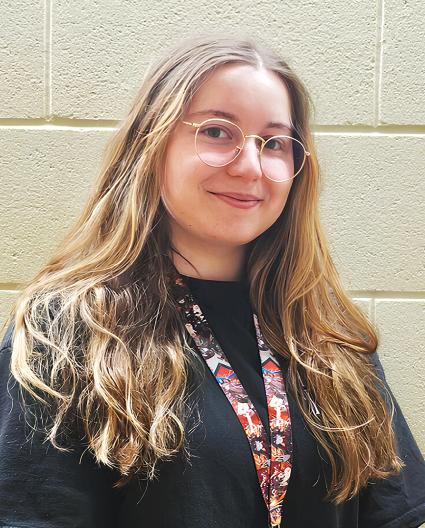 Une personne aux longs cheveux blonds portant des lunettes et un cordon porte-nom coloré sourit légèrement.