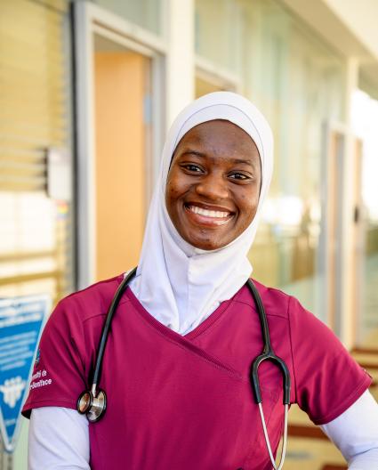 Une jeune femme portant un hidjab blanc, une tunique rouge vin d’infirmière et un stéthoscope sourit vers la caméra.