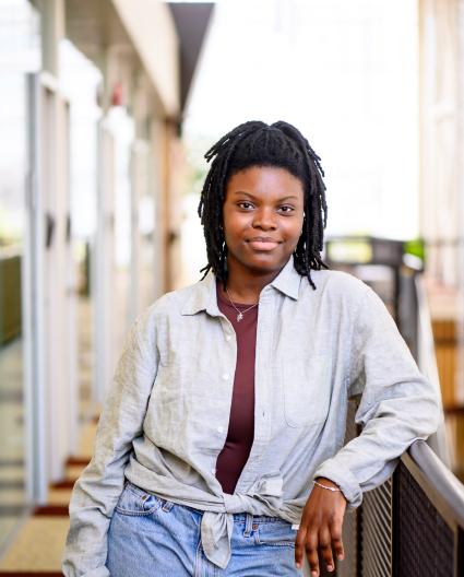 Une jeune femme aux cheveux noirs sourit à la caméra, alors qu'elle est adossée à la balustrade dans un couloir de l'université.