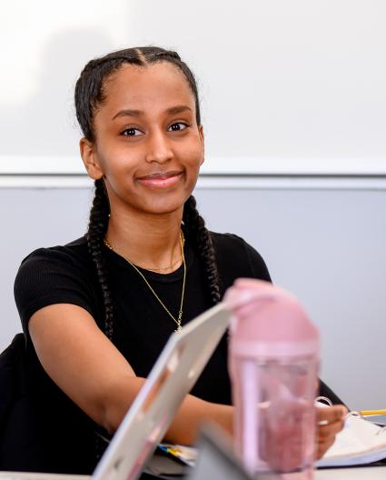 Une jeune femme aux cheveux noirs attachés sourit vers la caméra. Elle est en train de prendre des notes dans un cahier. 