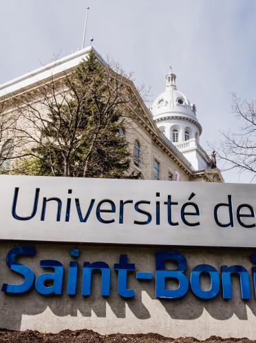 Monument d'entrée de l'Université de Saint-Boniface devant le bâtiment un jour de printemps, des arbres sont dépouillés de leurs feuilles.